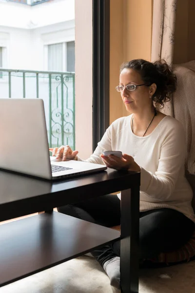 A woman sitting by a window in front of a laptop, telecommuting.
