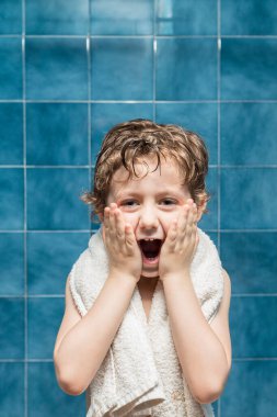 Omzunda havlu, yüzünde elleri, ağzı açık bir çocuk ve arkasında mavi fayanslar olan komik bir ifade..