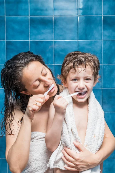 A woman and a child brushing their teeth with blue tiles in the background.