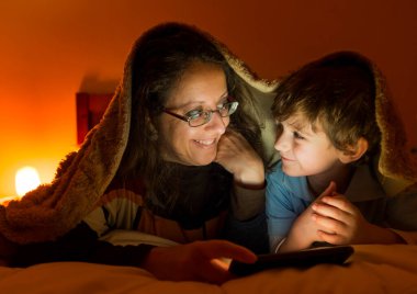 Bir çocuk ve bir kadın yatağa uzanıp battaniyeyle örtülmüş akıllı telefonu izliyorlar..