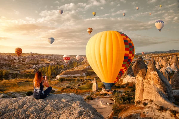 Jonge vrouw kijkt en fotografeert vliegende kleurrijke ballonnen op een vroege ochtend in Goreme Valley, Cappadocië. Turkije — Stockfoto