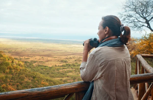 Vrouw staat op houten terras met camera kijken naar Afrikaanse savanne in de zomer vakantie. — Stockfoto
