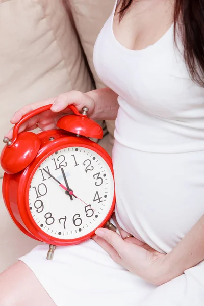 Pregnant woman sitting on sofa holding clock — Stock Photo, Image