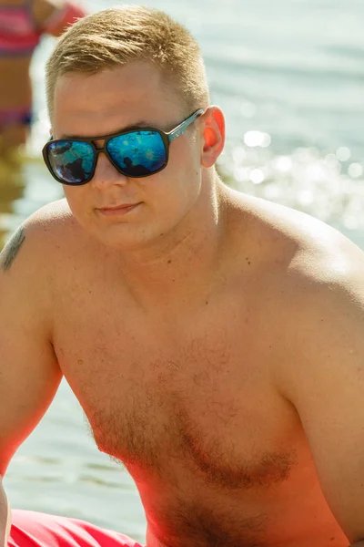 Deniz kenarında çömelmiş güneş gözlüklü adam — Stok fotoğraf