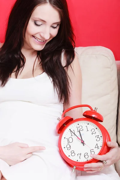 Pregnant woman sitting on sofa holding clock — Stock Photo, Image