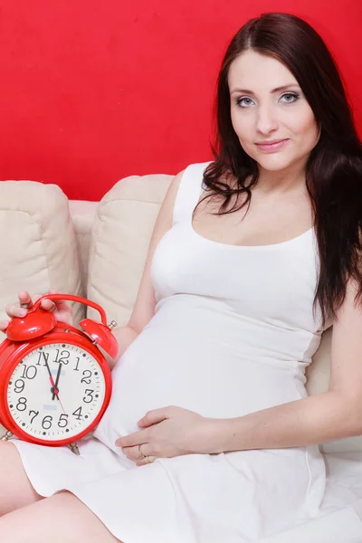 Zwangere vrouw zittend op de bank holding klok — Stockfoto