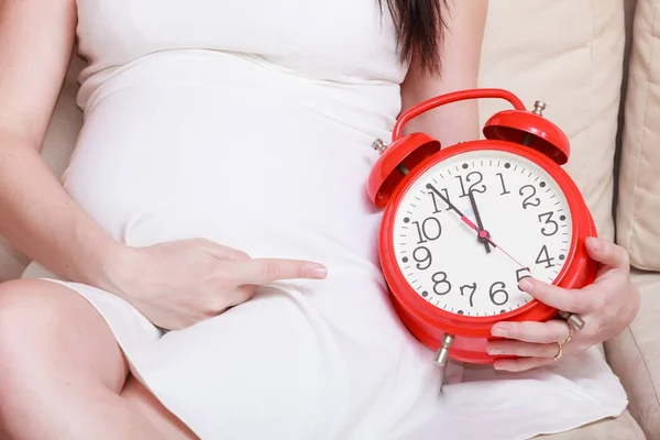 Pregnant woman sitting on sofa holding clock — Stock Photo, Image