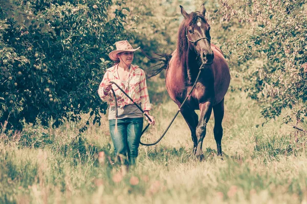 Δυτική γυναίκα το περπάτημα σε πράσινο λιβάδι με άλογο — Φωτογραφία Αρχείου