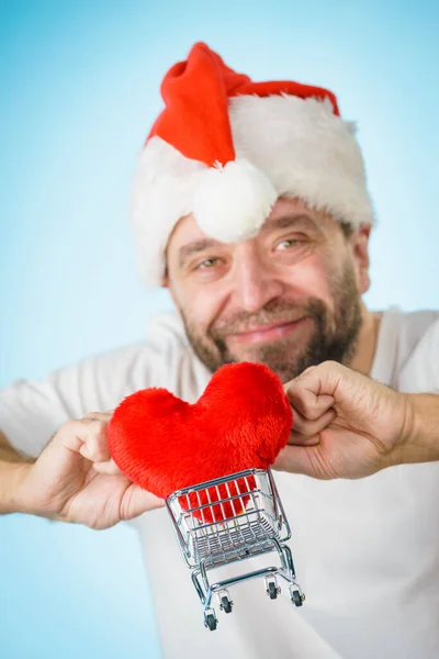 Mann mit Weihnachtsmütze hält Einkaufswagen mit Herz. — Stockfoto