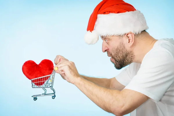 Mann mit Weihnachtsmütze hält Einkaufswagen mit Herz. — Stockfoto