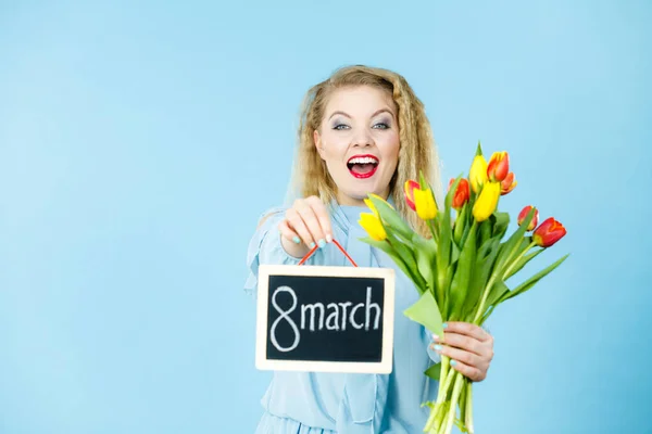 International women day. Beautiful woman blonde hair fashion make up holding red yellow tulips and frame board with message 8 march. On blue