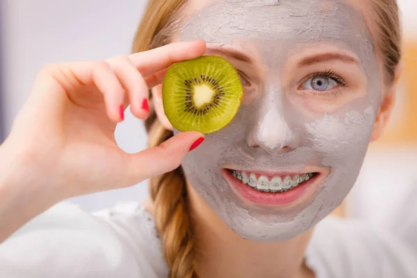 顔の乾燥肌と体のケア 肌治療ホーム コンセプトで キウイの部分を保持している彼女の顔に灰色の泥マスクを持っている幸せな若い女 — ストック写真