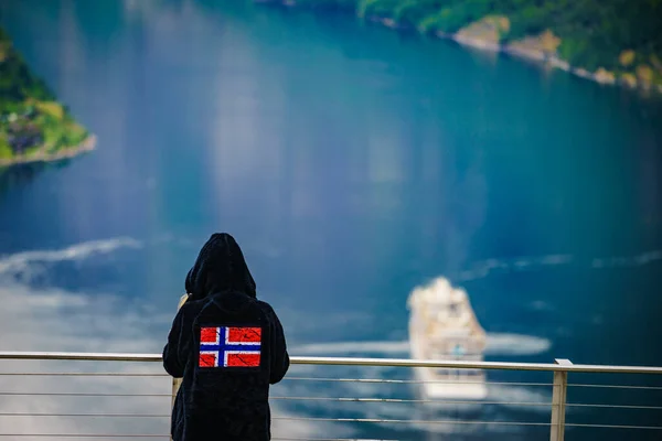 Tourist wearing norwegian flag clothing enjoying scenic view over fjord Geirangerfjorden from Flydalsjuvet viewpoint. Cruising vacation and travel.