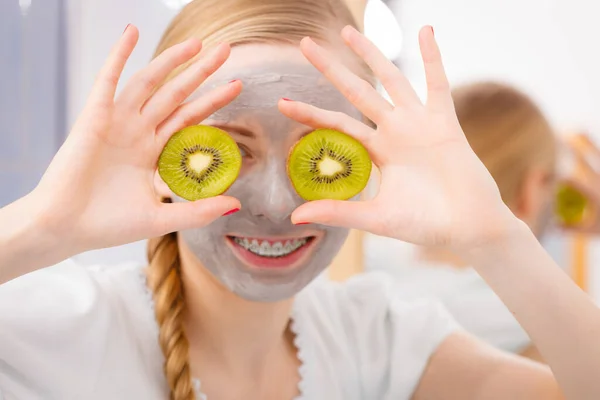 顔の乾燥肌と体のケア 肌治療ホーム コンセプトで 目のキウイの部分を保持している彼女の顔に灰色の泥マスクを持つ幸せな若い女 — ストック写真