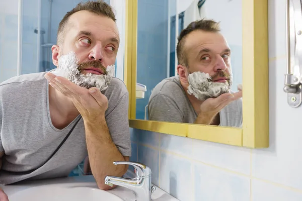 男の顔にシェービングフォームクリームを適用し バスルームに立って 鏡を見て 彼の顔ひげを剃る準備 スキンケア — ストック写真