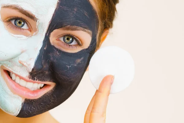 Woman with clay carbo black mask on one half face and white mud on second half. Girl removing cosmetic scrab with cotton swab. Female taking care of oily complexion. Skincare.