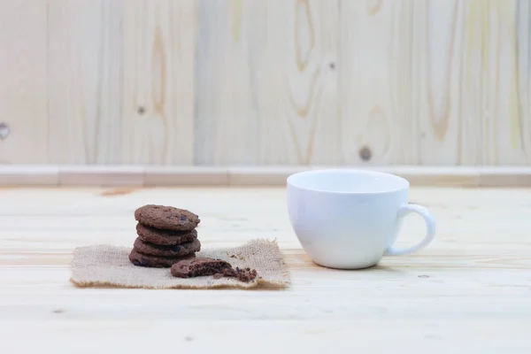 Chocolate chip cookies with a bite mark and a cup placed on a sack on a wooden table.