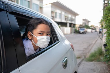 Küçük Asyalı kız kafasını arabanın camından dışarı çıkardı. Hijyen maskesi korumalı koronavirüs ya da kovid 19. Öğrenciler arabada oturur ve mikropları önlemek için maske takarlar..