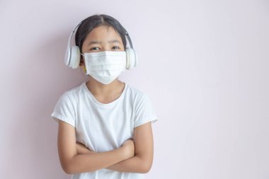 Tıbbi koruyucu maske ve kulaklık takan küçük bir Asyalı kız. Arka planda kolları çapraz duran kız çocuğu. Grip salgını, Coronavirüs veya COVID-19 ve hastalık konsepti.