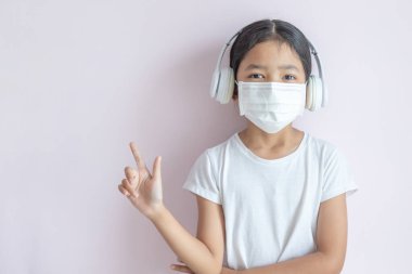 Tıbbi koruyucu maske takan ve sağ parmağını gösteren kulaklık takan küçük Asyalı kız. Grip salgını, Coronavirüs veya COVID-19 ve hastalık konsepti.