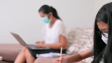 Coronavirüs veya COVID-19 konseptinin yayılmasını azaltmak için E-öğrenme ve sosyal mesafeler. Maskeli Asyalı kadınlar evde çalışıyor ve küçük bir kız evde okul için akıllı telefon kullanıyor..