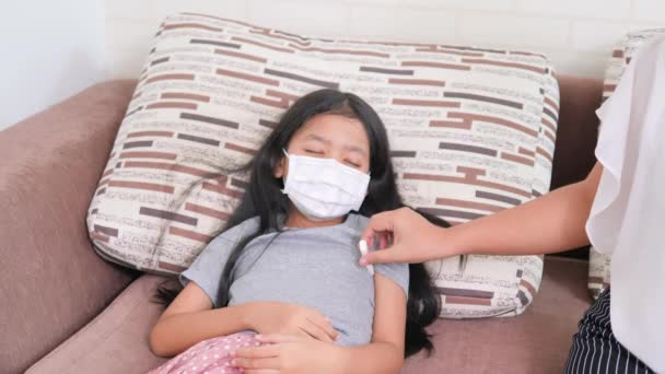 アジア系の少女は外科用マスクを着用し咳をしてソファの上に横になっている 女性は子供の温度計を使う — ストック動画