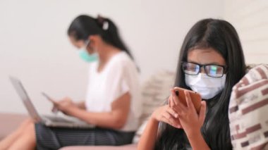Coronavirüs veya COVID-19 konseptinin yayılmasını azaltmak için E-öğrenme ve sosyal mesafeler. Maskeli Asyalı kadınlar evde çalışıyor ve küçük bir kız evde okul için akıllı telefon kullanıyor..