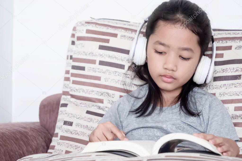 The little girl wore a white headset, sat on the couch and reading a book with intent.