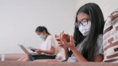 Küçük Asyalı kız kanepede oturmuş akıllı telefon çalarken gözlük ve maske takıyor. Evden çalışan ve kızından uzakta oturan bir kadın. Sosyal uzaklık kavramı için..