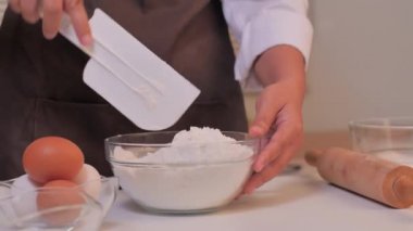 Video kaydırağı, kadınların eli beyaz bir spatula kullanarak temiz bir cam bardakta unu karıştırıyor.
