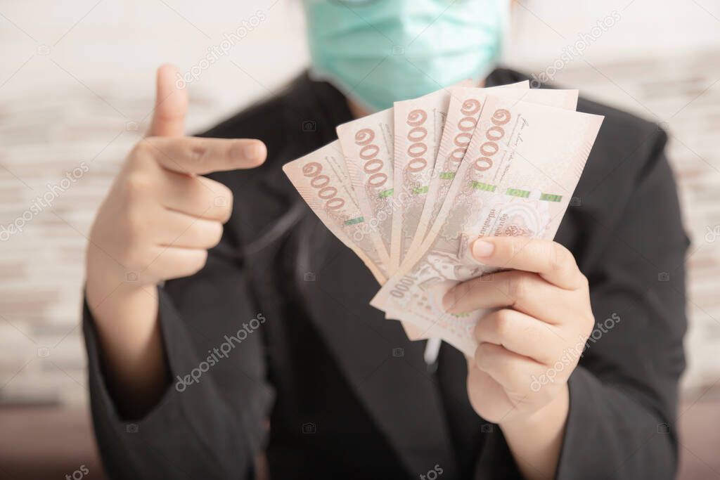 Hands of a woman holding a banknotes total amount of 5,000 baht of Thailand. A human wearing a medical mask and a black suit pointing the money in her hand.