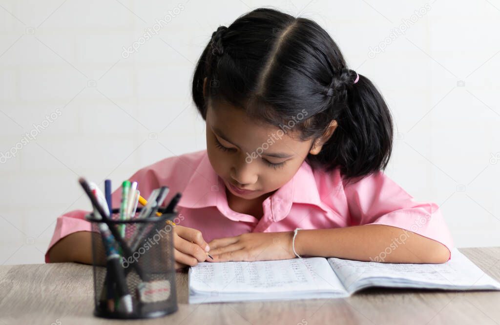 The little girl is doing homework intently. Children use a yellow pencil is writing a notebook on the wooden table. Select focus shallow depth of field with copy space on white background.