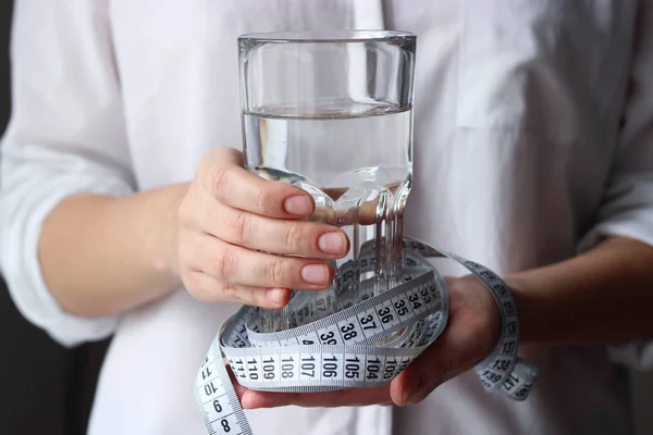a glass of water and a centimeter tape in female hands on a light background. use of water. slimming, minimalism, health, diet, proper nutrition.