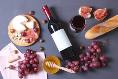 Masanın üstünde kırmızı şarap ve atıştırmalıklar var. Şarap, prosciutto, peynir, baget, üzüm, bal.