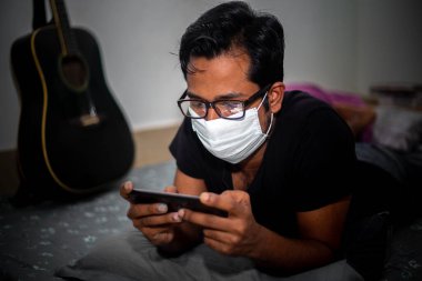 Cerrahi maske takan genç bir adam, Dhaka 'daki koronavirüs ev karantinası nedeniyle kendi evinde mobil oyunlar oynuyordu..