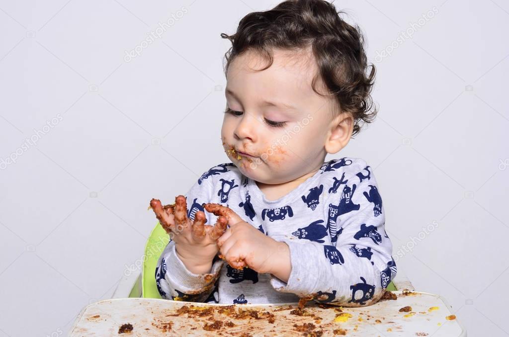 One year old kid eating a slice of birthday smash cake by himself getting dirty.