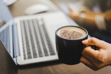 Laptop 'ta çalışan ve kahve bardağını elinde tutan genç bir kadın..