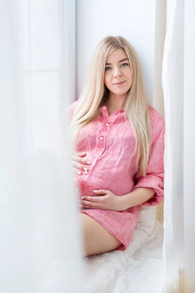 Schöne schwangere Blondine posiert am Fenster für die Kamera — Stockfoto
