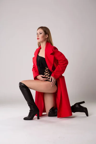 一个年轻迷人的女模特 身穿红色雨衣 身穿黑色性感紧身衣 身材苗条 坐在白色孤立的背景下 工作室肖像 — 图库照片