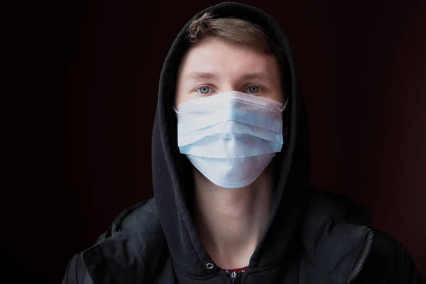 A young man in black clothing with a hood on his head and a medical mask on his face against the coronavirus, looks into the camera. Concept of the quarantine period of the ncov-19 coronavirus pandemic.