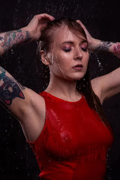 一位穿着红色湿T恤的性感金发姑娘紧闭双眼 站在鲜亮的水花下 — 图库照片