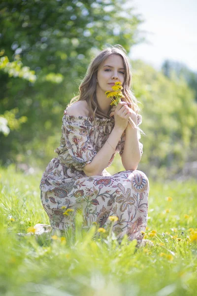 A beautiful blonde girl in a summer light dress inhales the pleasant aroma of the first flowers