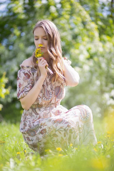 在公园草坪上 一位身穿独创浅色连衣裙的年轻漂亮女子的画像 她吸入着鲜花的芬芳 — 图库照片
