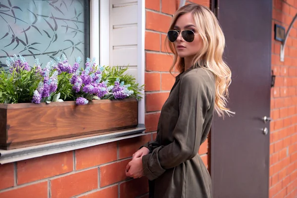 Une belle blonde aux lunettes élégantes se tient à la fenêtre d'une maison décorée de fleurs de la rue — Photo