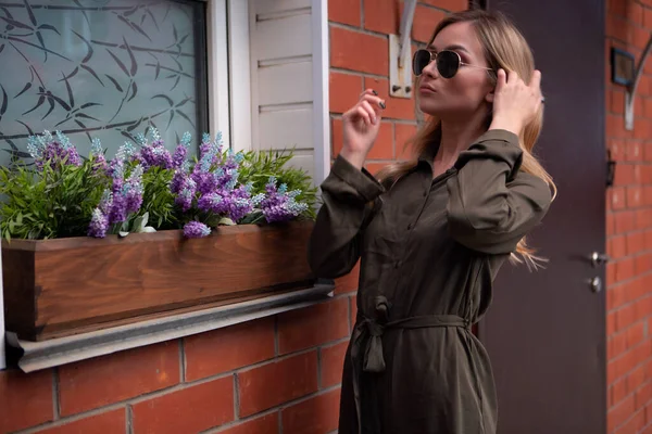 Une belle blonde aux lunettes élégantes se tient à la fenêtre d'une maison décorée de fleurs de la rue — Photo