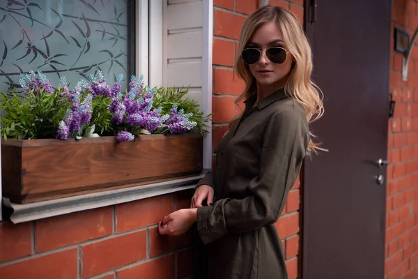 Uma linda garota loira em óculos elegantes fica na janela de uma casa decorada com flores da rua — Fotografia de Stock