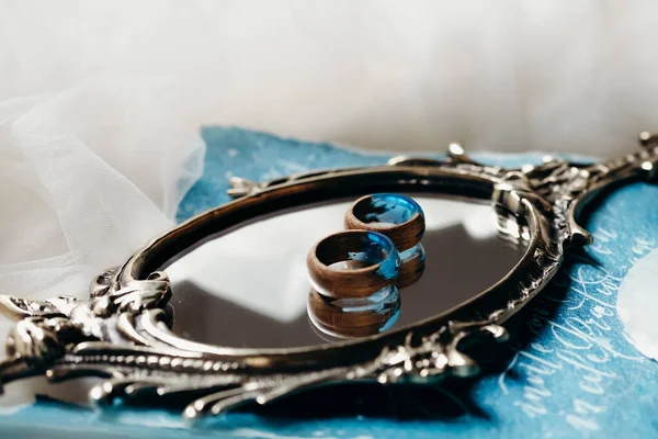 镜子上挂着环氧树脂蓝色的木制戒指 蓝色的婚宴请柬 还有特写的郁金香织物 — 图库照片