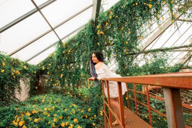 Beyaz elbiseli, çok renkli rastalı yalnız, güzel bir kız kırmızı bir merdivende, bir çiçek kemerinde duruyor. Ukrayna, Dnipr 'deki botanik bahçesinde egzotik tropik bitkilerle dolu bir sera.