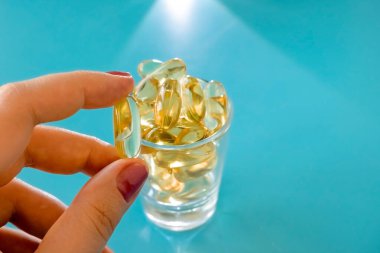 Kadın eli saydam bir omega 3 kapsülü, mavi bir masanın üzerinde haplar olan bir bardak tutuyor.