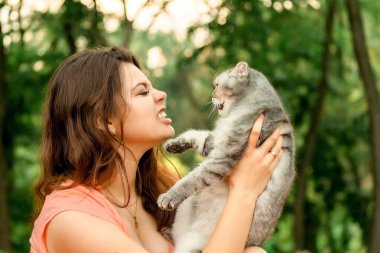 Parkta kedisiyle oturan güzel, uzun saçlı esmer kız. Kız kediye hırlıyor ve ısırıyor. Sokakta evcil bir kediyle fotoğraf çektirmek.